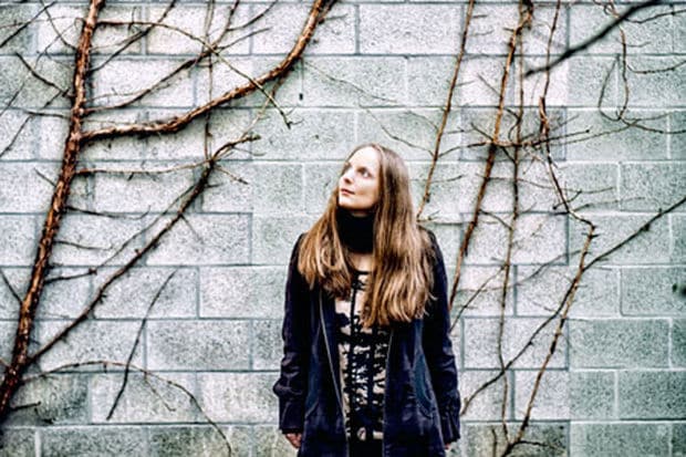 Karin Bernfeld adossée à un mur de pierre, sous des branches nues, le regard levé