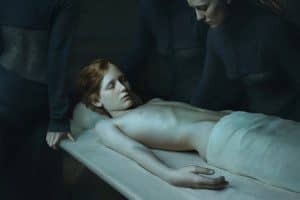 Photographie de Désirée Dorlon : une jeune femme au corps décharné allongée sur une table, veillée par des silhouettes vêtues de sombre